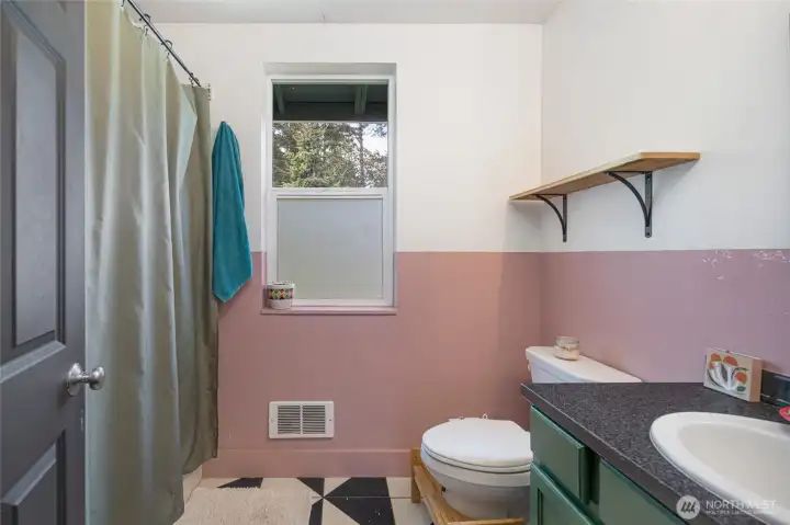 This thoughtfully designed bathroom makes a lasting impression with its graphic black-and-white geometric tile floor, dusty rose wainscoting, sage green cabinetry, and natural light streaming through a frosted privacy window — a warm, artistic space that feels personally crafted rather than builder-grade.