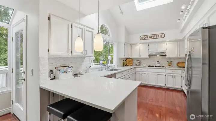 The kitchen is bright and open with white quartz counters white stacked tile backsplash, custom lighting and a large sky light as part of the vaulted ceiling. There is a garden window with a half round window above.  All the appliances remain with the home and all are Stainless steel.