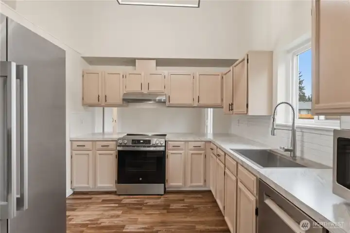 Bright and efficient kitchen with generous cabinetry, LVP flooring, and stainless steel appliances, highlighted by a window above the sink and an open bar to serve through.