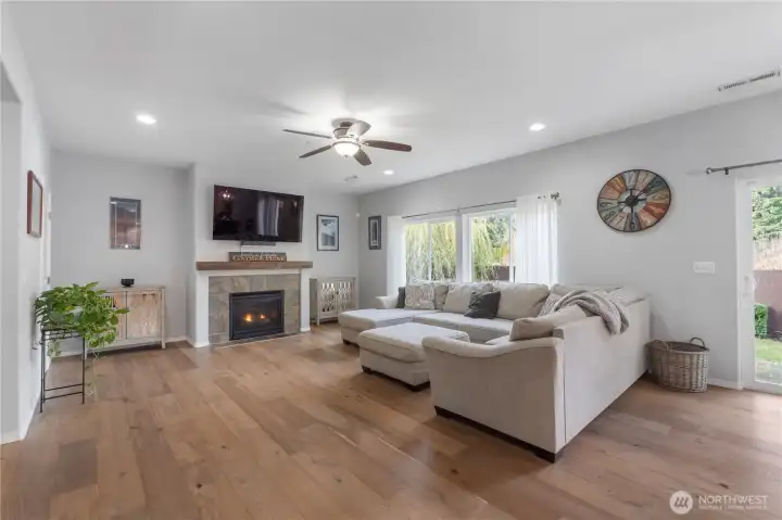 A comfortable living room centered around a gas fireplace with a stone surround and wood mantel. Large windows bring in soft natural light, while the layout easily accommodates a sectional sofa. This is a warm, inviting place to relax at the heart of the home.
