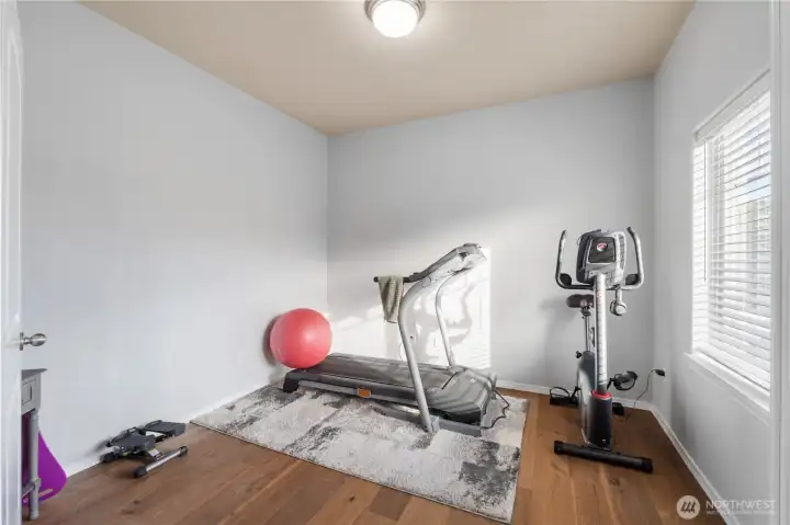 A bright flex room set up as a home gym, featuring a treadmill, stationary bike, and natural light from a large window. The soft wall color and warm hardwood floors create a calm, focused environment that can easily adapt to a home office, playroom, or guest space.
