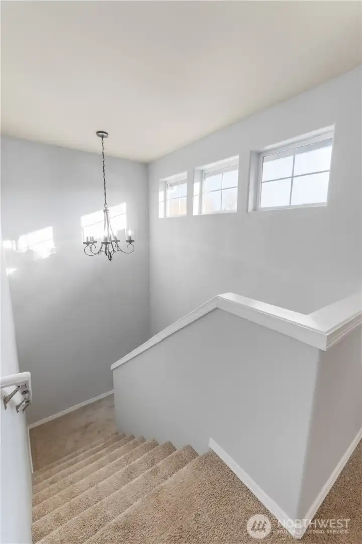 Light pours into this bright stairwell through a row of transom-style windows, highlighting clean lines, soft carpet underfoot, and a stylish hanging chandelier. It gives the entry a welcoming, open feel.