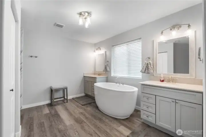 A spacious, updated primary bathroom featuring dual vanities, a sleek freestanding soaking tub, and a large window that fills the room with natural light. Soft gray cabinetry and modern lighting create a calm, spa-like atmosphere.