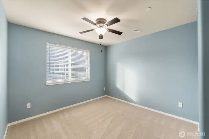 A bright guest or secondary bedroom with soft blue walls and warm natural light. Clean carpet, a ceiling fan, and simple lines create a calm, comfortable space ready for personal touches.