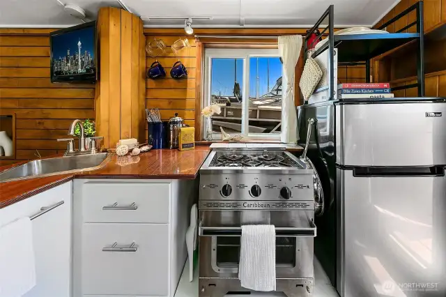 The galley’s warm wood cabinetry frames a propane Dickinson Marine Caribbean 2-burner stove/oven, efficient for daily cooking with a single 5-gallon tank lasting months — all set against the backdrop of Lake Union.