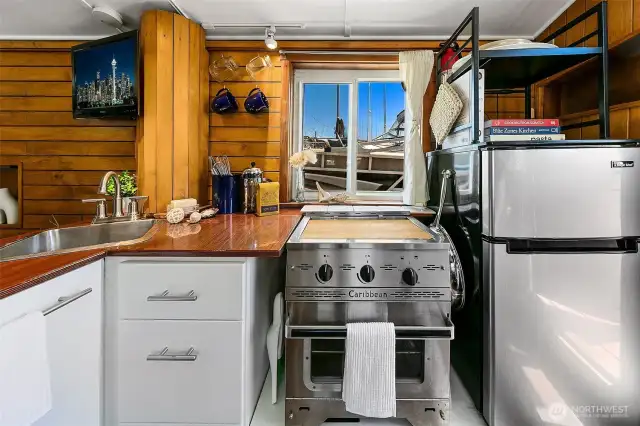 The galley’s warm wood cabinetry frames a propane Dickinson Marine Caribbean 2-burner stove/oven, efficient for daily cooking with a single 5-gallon tank lasting months — all set against the backdrop of Lake Union.