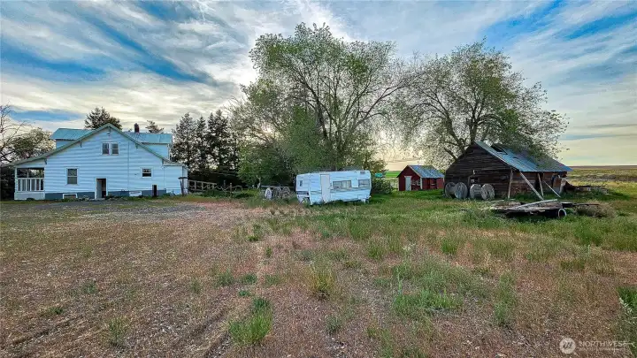 West-facing view of the home's east-side exterior, highlighting the expansive parking area, mature trees, and multiple outbuildings.