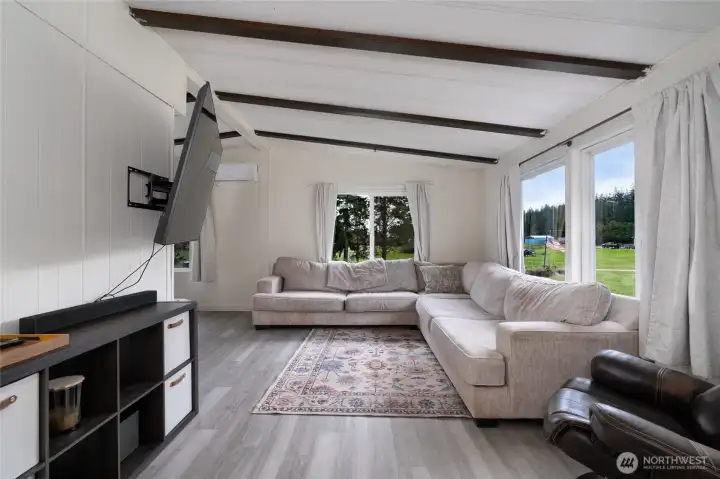 Comfortable living room with large windows offering peaceful views of the surrounding landscape. Features include vaulted ceilings with exposed beams, modern flooring, and plenty of natural light—ideal for relaxing or hosting guests.