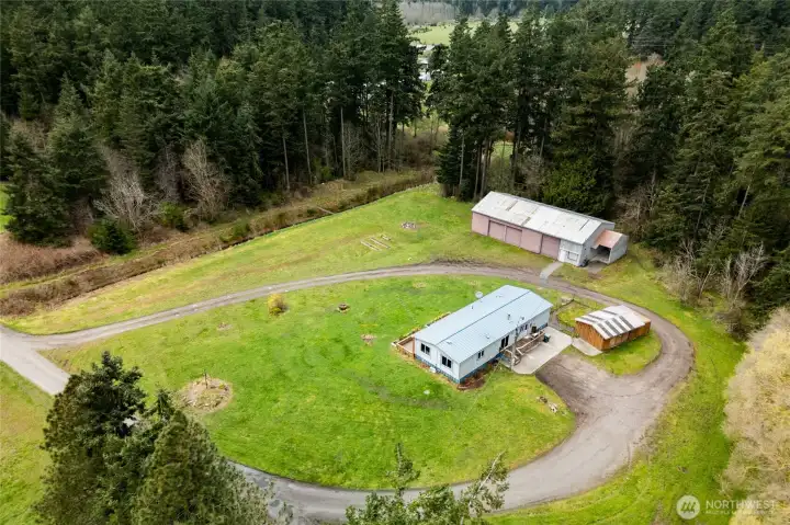 Scenic aerial view of a rural property featuring a single-level home, large detached shop/garage, and expansive usable acreage. A curved driveway provides easy access, while the surrounding trees offer privacy and a peaceful country setting—ideal for those seeking space, functionality, and tranquility.