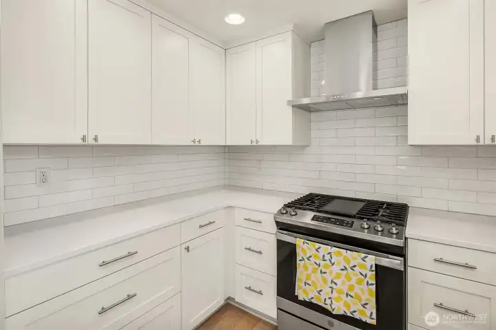 Designer kitchen featuring white shaker cabinetry, quartz countertops, full-height subway tile backsplash, stainless steel range hood, gas range with griddle, and abundant cabinet storage
