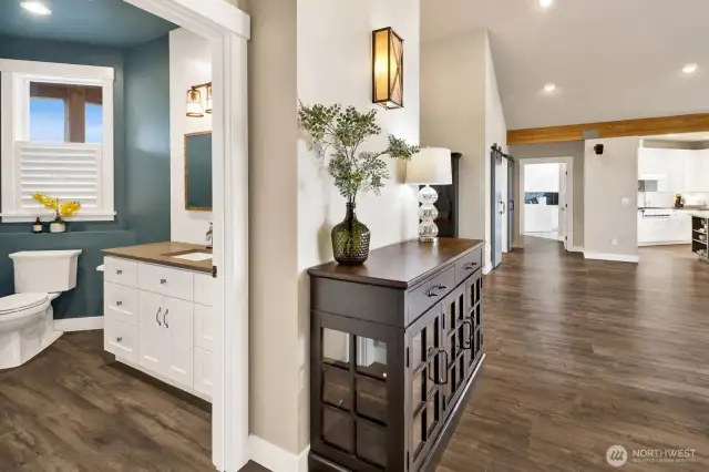 Views of half bathroom and main living area. Off in the distance is a small glimpse of the kitchen and straight ahead is the large laundry/mudroom leading out to the garage.
