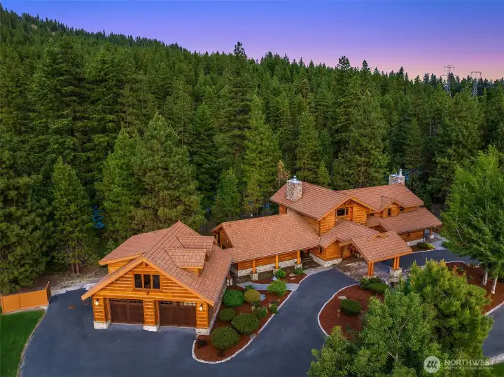 Elevated aerial perspective showcasing the estate’s striking lodge architecture, thoughtfully positioned within a blanket of mature pines. A sweeping, paved driveway leads to the expansive detached garage and covered entry, creating a true sense of arrival and privacy.