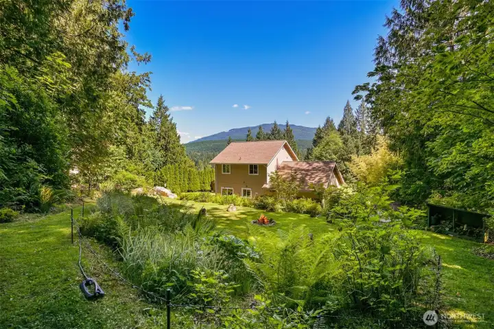From the upper lawn and fire pit area, the home sits within a sunny clearing surrounded by forest, gardens, and distant mountain views. This perspective helps illustrate the property's unique combination of open usable space and natural privacy. From the upper lawn and fire pit area, the home sits within a sunny clearing surrounded by forest, gardens, and distant mountain views. This perspective helps illustrate the property's unique combination of open usable space and natural privacy.
