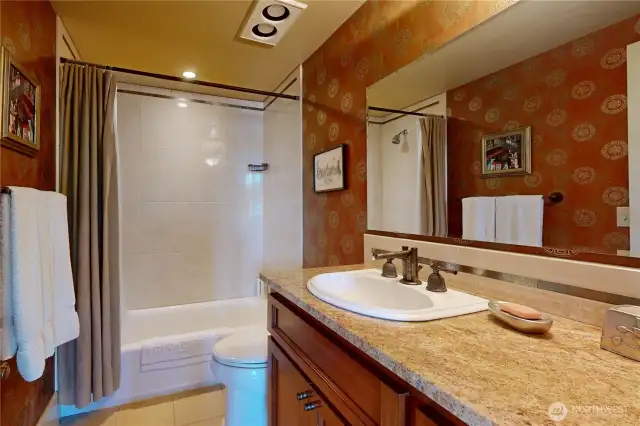 Guest bathroom – granite countertop, tiled shower/tub combo, and warm-toned wallpaper accent.