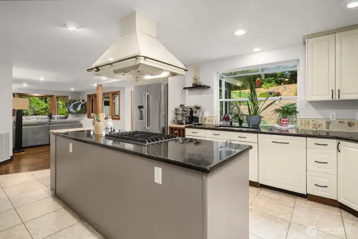 This view of the kitchen showcases the oversized island with dramatic dark counters, a statement range hood, and an abundance of windows that bathe the space in natural light and frame the gorgeous outdoor views.