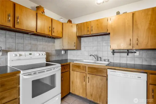 This kitchen on the lower floor has plenty of cupboard space, pull-out drawers, all white appliances (stove/range, dishwasher, and fridge), tile flooring and light-gray tile backsplash that goes all the way up to the cupboards. If you need extra storage, there's plenty of room on top of the cupboards where the cookie jar collection currently is.