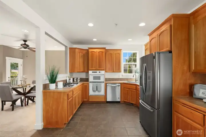 The kitchen on main level  includes a Frigidaire 5-burner gas cooktop, KitchenAid microwave, built-in oven, Samsung stainless steel refrigerator, large single-basin stainless steel sink, abundant cabinetry, and backyard views.