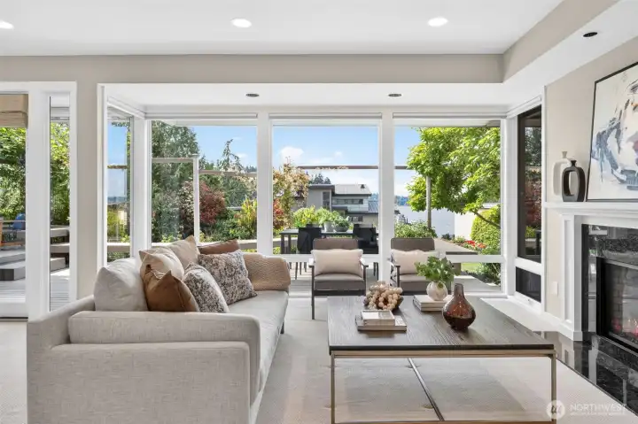 This is the shot that sells the home. Floor-to-ceiling new windows frame a direct view of Lake Washington and the lush Ohashi-designed back garden, blurring the line between the living room and the Pacific Northwest landscape beyond. The gas log fireplace glows to the right while the deck and outdoor seating beckon just past the glass — a home that lives as beautifully outside as it does in.