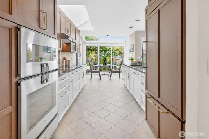 A chef's kitchen built for performance — Gaggenau wall ovens, custom Acorn cabinetry in rich walnut and crisp white, and dual skylights that bathe the entire galley in natural light. The kitchen terminates in a glass breakfast room framed by the pergola garden, turning every morning coffee into a moment worth savoring.