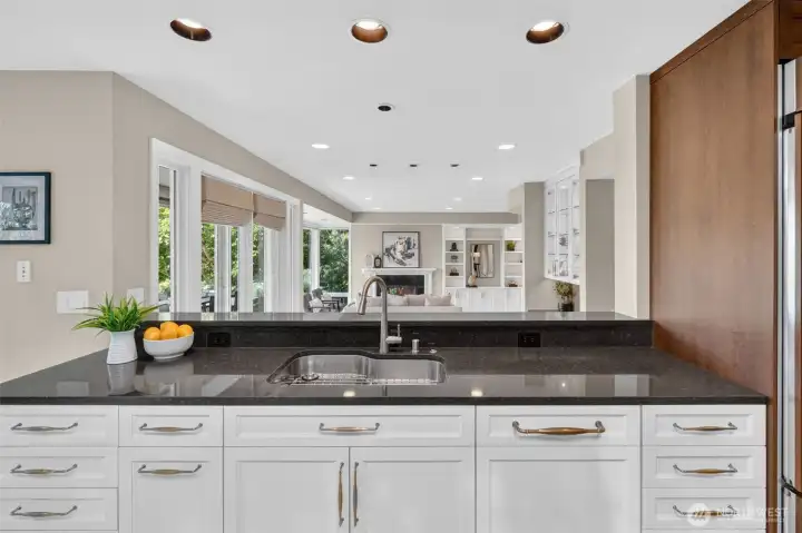 Dark granite countertops, brushed gold hardware, and an undermount sink anchor this working peninsula, while the pass-through opens directly to the living room and fireplace beyond — keeping the cook connected to every conversation. The scale of counter space and cabinetry here reflects a kitchen designed for someone who actually uses one.