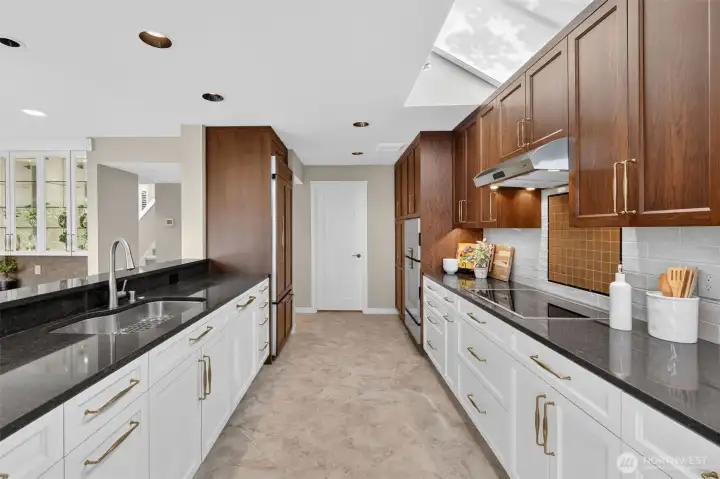 The full galley stretch reveals just how much storage and prep space this kitchen truly delivers — dual granite runs, gold hardware, and a Gaggenau induction cooktop on one side, undermount sink and panel-ready refrigerator on the other. The door at the end leads directly to the mudroom and garage, keeping arrivals and departures seamlessly tucked away from the main living areas.