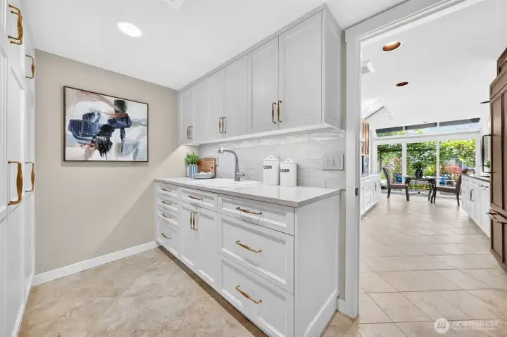 A mudroom that refuses to look like one — custom cabinetry to the ceiling, quartz counters, gold hardware, a utility sink, and subway tile backsplash give this hardworking space the same fit and finish as the rest of the home. The glass breakfast nook and garden room visible straight ahead make even the daily drop zone feel light and connected.