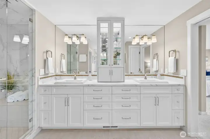 The dual vanity wall is the centerpiece of the ensuite — two undermount sinks in Calacatta Dekton, a glass-front illuminated linen tower anchoring the center, and matching mirror panels stretching the full width. Polished nickel hardware adds warmth against the crisp white cabinetry, while the steam shower visible to the left reminds you of the full scope of this space. This is a bathroom designed for two people who never have to compromise. Notice the appliance garage for all your hair tools.