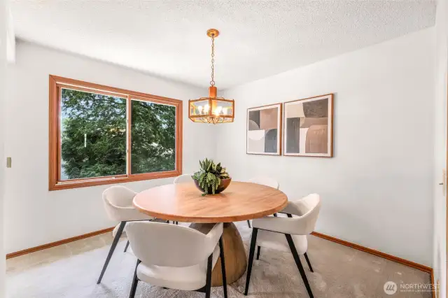 Light-filled dining area featuring a stylish pendant light, contemporary round wood table, and plush seating, framed by large windows that offer peaceful views of lush greenery. Neutral tones and modern wall art add a sophisticated touch to this versatile space.