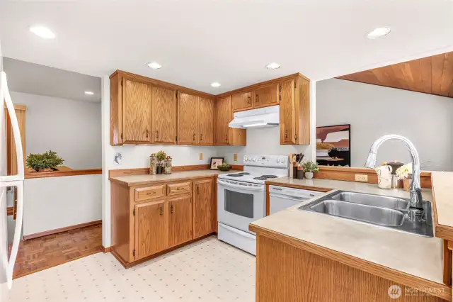 Expanded view of the kitchen reveals a spacious design with peninsula counter, abundant cabinet storage, and open sight lines to the dining and living areas. A large window over the sink enhances natural lighting.
