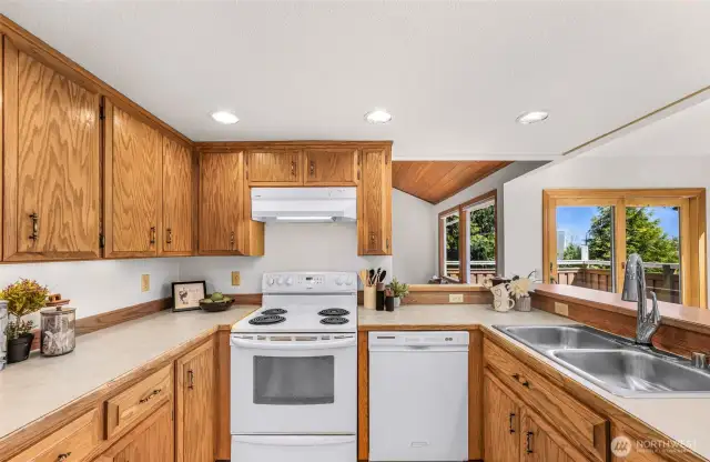 This kitchen features warm oak cabinetry, white appliances, and a U-shaped layout with ample counter space. Recessed lighting and a pass-through window create a bright, connected feel to the adjoining rooms.