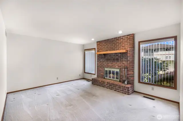 Spacious living room with a statement brick fireplace and wood mantel, flanked by large windows with vertical blinds that allow for privacy and light control. Clean carpet and recessed lighting complete the space.