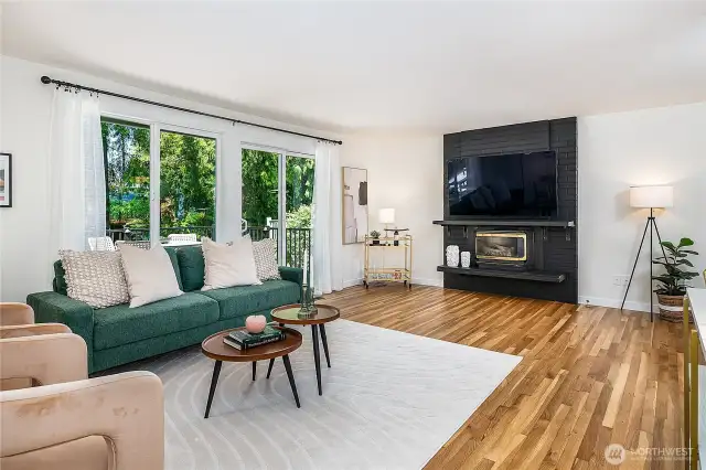 Bright and inviting main-level living room featuring a classic brick fireplace with gas insert, abundant windows, and a slider that opens to a spacious deck.
