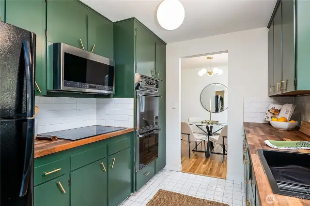 Main kitchen with freshly painted forest green cabinets, new cooktop, butcher block countertops, updated sink, tile backsplash, and modern hardware. Opens to the dining room for easy flow.