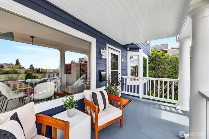 Welcoming covered front porch with rich blue siding, classic columns, cozy seating areas, and a bright indoor-outdoor connection perfect for relaxing.