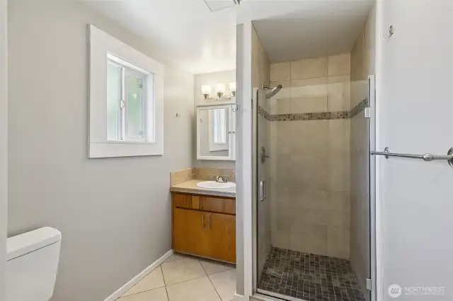 Unit A’s 3/4 bathroom, attached to the back Bedroom #1, creates the perfect primary en suite. The glass door tile shower adds a sleek touch, and the medicine cabinet above the sink offers convenient storage. A window that opens provides fresh air, while the tile floors and light grey walls with white trim complete the modern, clean look.