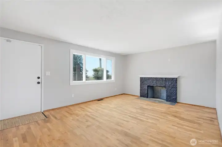 Spacious living room featuring hardwood floors, a classic wood-burning fireplace, and large picture windows that bring in abundant natural light. A welcoming space for gathering, relaxing, and everyday living.