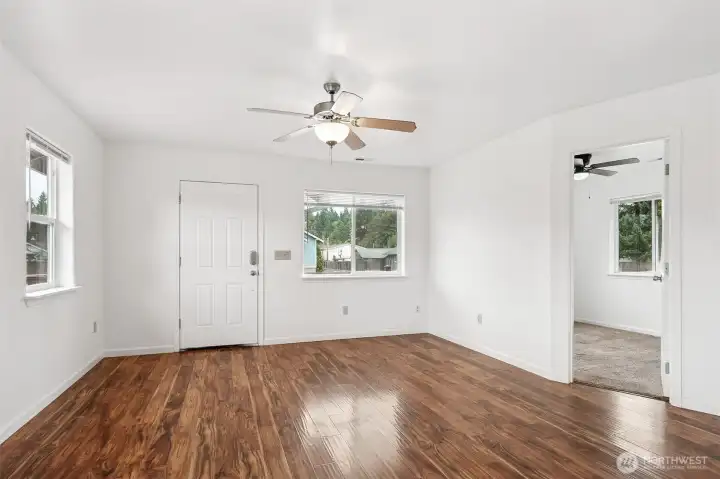 This living room makes a strong first impression with warm wood-tone luxury vinyl plank flooring that can handle pets, kids, or the upcoming-wet-weather-life we call home in the PNW. The white walls are a blank slate, ready for any design personality you bring in, whether you’re into high-contrast drama or quiet neutrals. A ceiling fan, front and side windows, and an open view into one of the five bedrooms keep the space light and flexible.