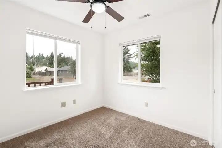 The front bedroom gets the corner treatment with twin windows and blinds, so there’s plenty of light without sacrificing privacy. White walls and latte-toned carpet keep it neutral, while the ceiling fan and ceiling-mounted heat register prove someone was actually thinking about comfort and furniture placement.