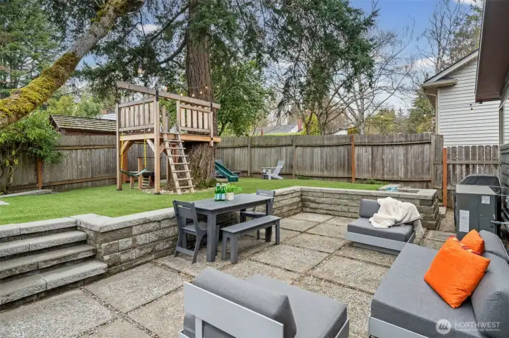 A sun-drenched entertaining terrace designed for effortless gatherings, with structured stonework and defined outdoor living zones. The elevated turf lawn creates a lush, low-maintenance green backdrop framed by mature evergreens, offering year-round beauty and functionality.