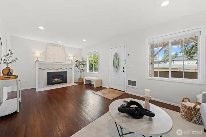The living room centers around a classic tile-surround propane fireplace, creating a warm and inviting anchor for the space. Rich hardwood floors, large windows on two sides, and recessed lighting make this room bright and comfortable year-round.