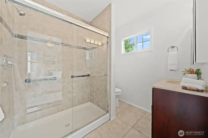 The ensuite bath off Primary Suite 1 features a generous walk-in shower with full-height ceramic tile surround, a sleek sliding glass enclosure, and a built-in mosaic accent strip that adds a polished finishing touch. A rich cherry vanity with quartz countertop, ceramic tile flooring, and a bright operable window round out this thoughtfully designed private bath. Everything here is clean, fresh, and move-in ready.