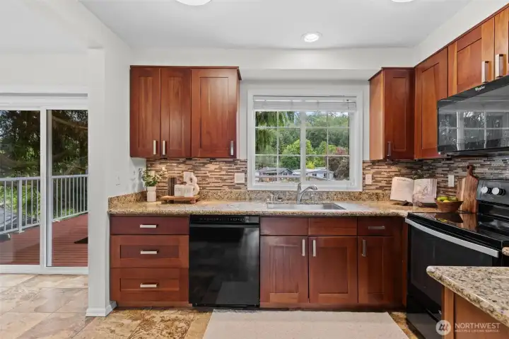 This kitchen features rich cherry shaker cabinetry, granite countertops, a stone mosaic tile backsplash, and a slide-to-open window above the sink allows for easy pass through when entertaining. Gaze out to views of the riverfront and lush greenery. The layout is functional and inviting, with a direct connection to the breakfast nook and outdoor deck for seamless indoor-outdoor flow.
