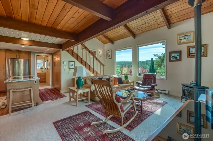 Main house living room with freestanding propane stove. Big windows looking west, vaulted ceiling and stairs to the upper two bedrooms/full bath.