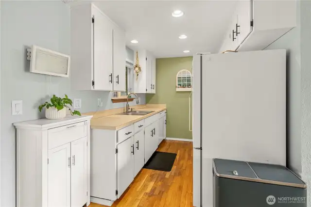 A bright kitchen with updated white cabinetry and black hardware, highlighted by hardwood floors and recessed lighting. A large sink and ample counter space provide functionality, while a window above the sink offers backyard views and natural light.
