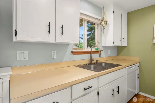 Close-up of the sink and prep area beneath the window. White upper and lower cabinets and simple laminate countertops offer easy-to-maintain surfaces with a timeless feel.