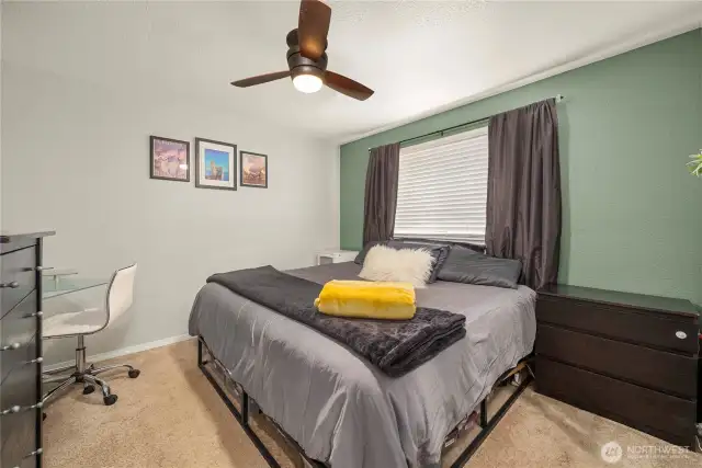 This spacious primary suite features ample room for a king-sized bed and additional storage options. Natural light pours in through the large window, highlighting the soft neutral palette and a calming green accent wall. Overhead, a modern ceiling fan adds both comfort and style.