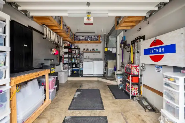 This well-maintained single-car garage offers both secure parking and ample workspace. Overhead and wall-mounted shelving provide smart storage solutions, while pegboard walls and open shelving make organization easy. At the rear, a dedicated laundry area features a side-by-side washer and dryer, a folding shelf, and utility hookups, all conveniently located near the home’s mechanical systems.