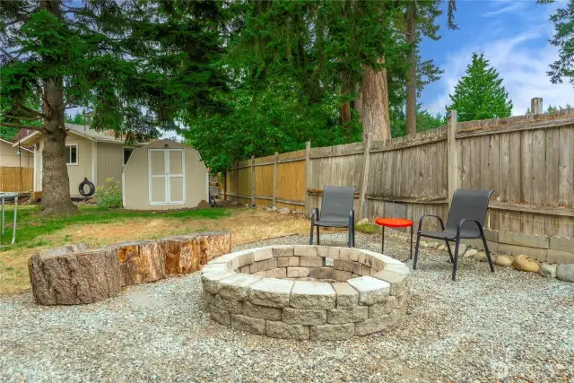 Cozy gravel fire pit area with a built-in stone ring, log-style seating, and additional chairs—perfect for gathering under the trees.