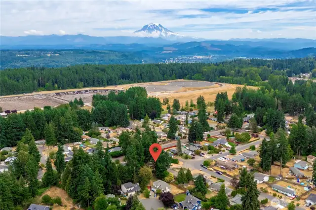 This elevated aerial image captures the home's setting within a peaceful, tree-lined neighborhood with a stunning backdrop of Mount Rainier. The property is marked with a location pin for easy identification, emphasizing its proximity to nature and the serene Pacific Northwest surroundings.