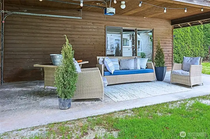 A versatile covered outdoor space extending from the detached garage, featuring one enclosed bay and a second open bay styled as a cozy lounge. With string lighting overhead and ample room for seating, it’s perfect for entertaining, relaxing, or creating a flexible workspace. A versatile covered outdoor space extending from the detached garage, featuring one enclosed bay and a second open bay styled as a cozy lounge. With string lighting overhead and ample room for seating, it’s perfect for entertaining, relaxing, or creating a flexible workspace.